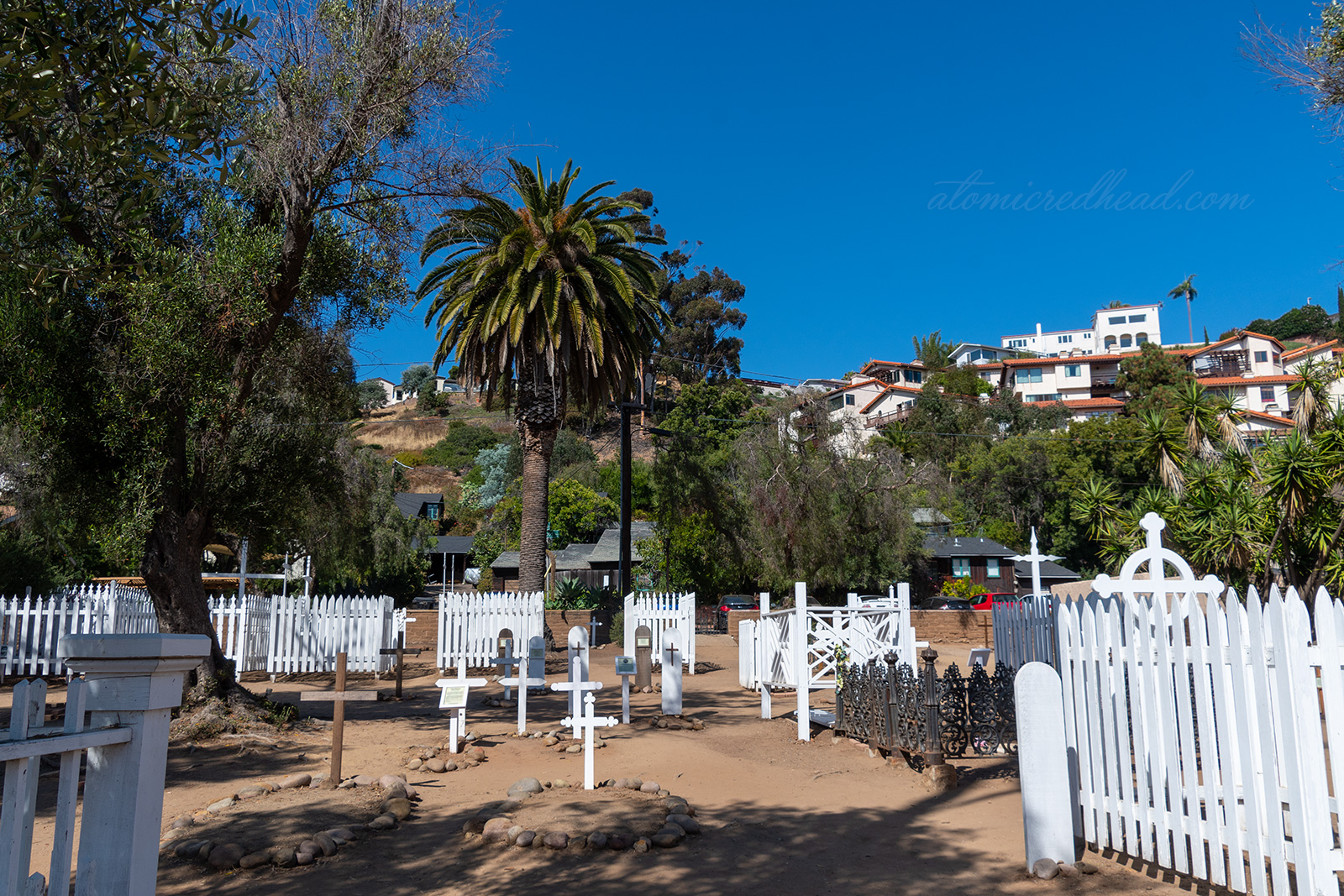 White painted wooden crosses mark the graves of some of San Diego's early residents. Some graves have white picket fences around them.