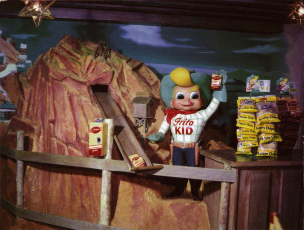 A large, cartoon looking cowboy in a blue hat atop blonde hair, wearing a red bandana around his neck, a white shirt with blue plaid and red text reading "Frito Kid" holding a bag of Fritos stands next to a mountain with a long slide coming from the top.