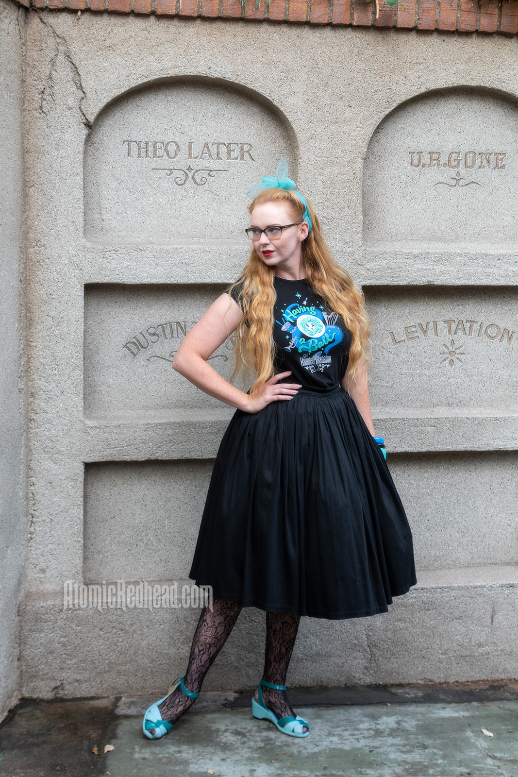 Myself standing in front of a faux mausoleum at the Haunted Mansion, wearing a black t-shirt with Madam Leota (a medium with her head floating in a crystal ball) and music instruments floating about on it, reading "Having a Ball" and a black full skirt.