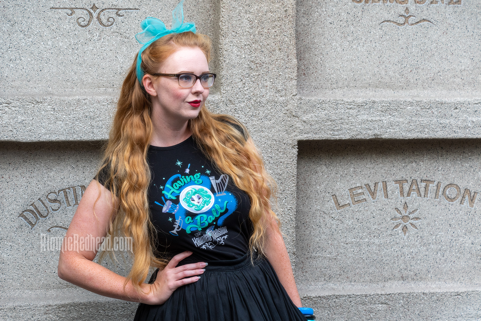 Myself standing in front of a faux mausoleum at the Haunted Mansion, wearing a black t-shirt with Madam Leota (a medium with her head floating in a crystal ball) and music instruments floating about on it, reading "Having a Ball" and a black full skirt.