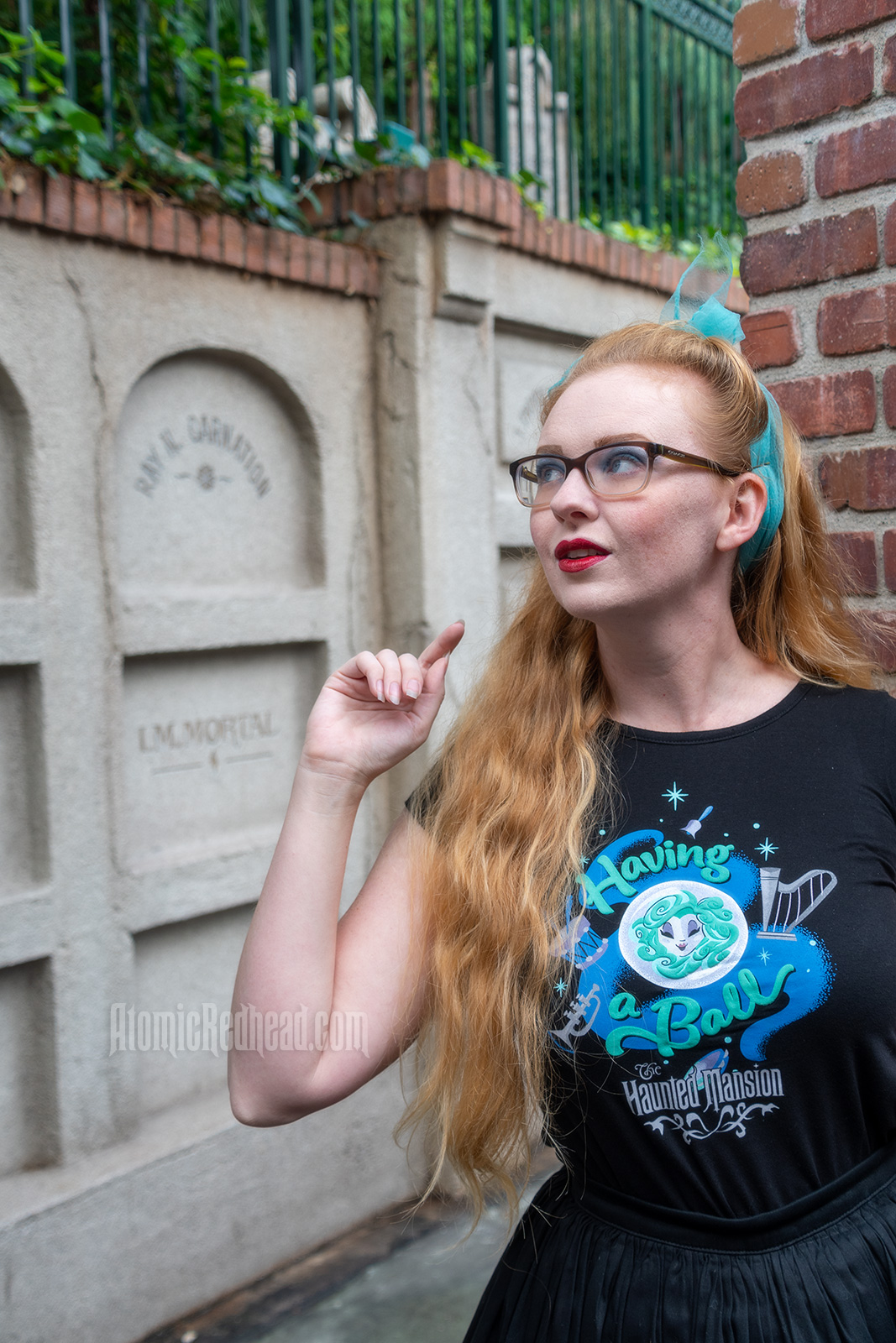 Myself standing in front of a faux mausoleum at the Haunted Mansion, wearing a black t-shirt with Madam Leota (a medium with her head floating in a crystal ball) and music instruments floating about on it, reading "Having a Ball" and a black full skirt.