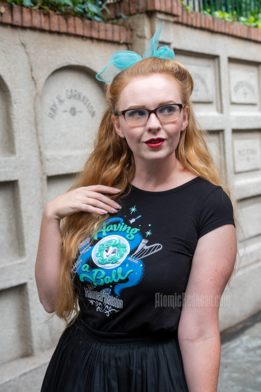 Myself standing in front of a faux mausoleum at the Haunted Mansion, wearing a black t-shirt with Madam Leota (a medium with her head floating in a crystal ball) and music instruments floating about on it, reading "Having a Ball" and a black full skirt.