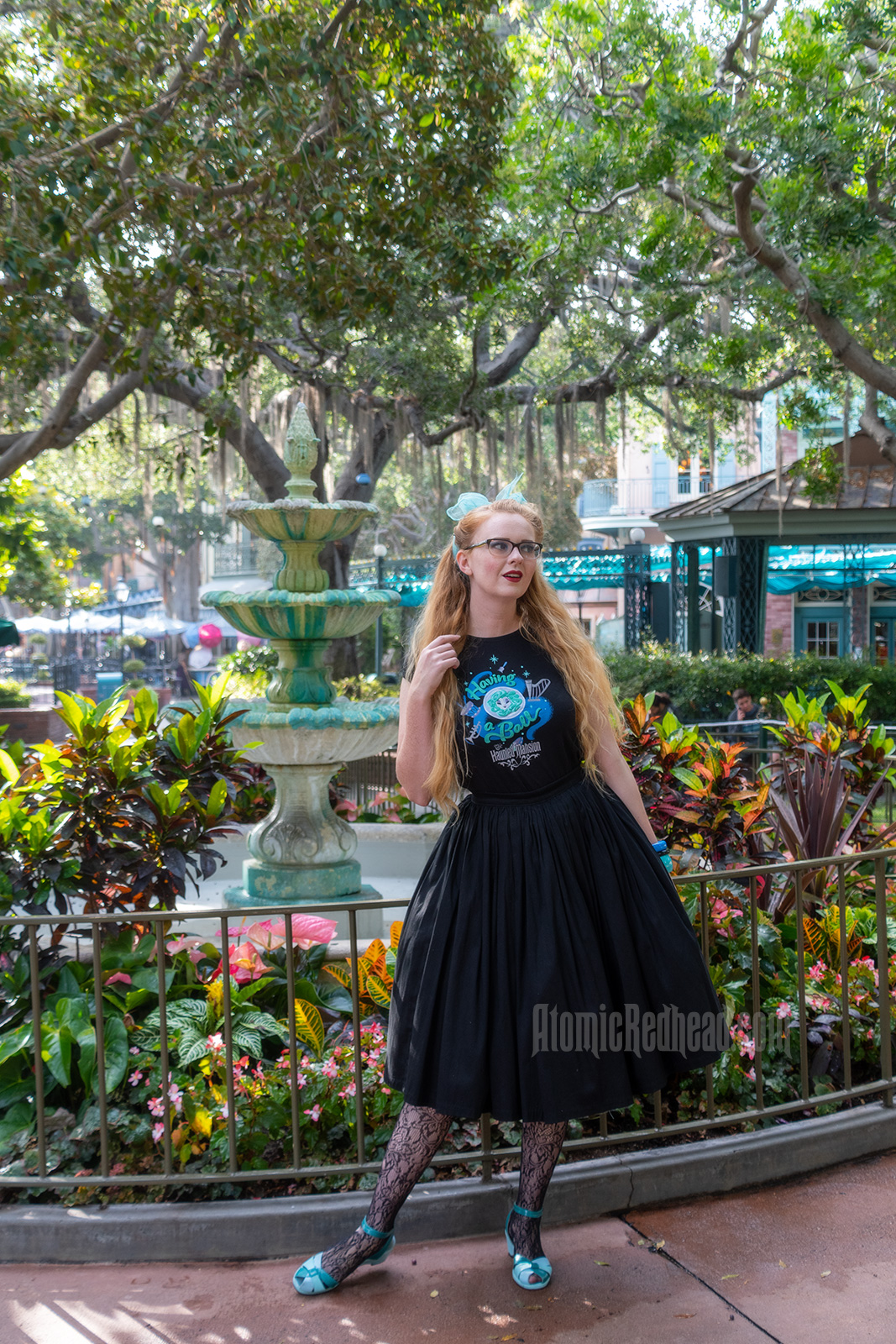 Myself standing in front of a fountain surrounded by trees dripping in Spanish moss, wearing a black t-shirt with Madam Leota (a medium with her head floating in a crystal ball) and music instruments floating about on it, reading "Having a Ball" and a black full skirt.