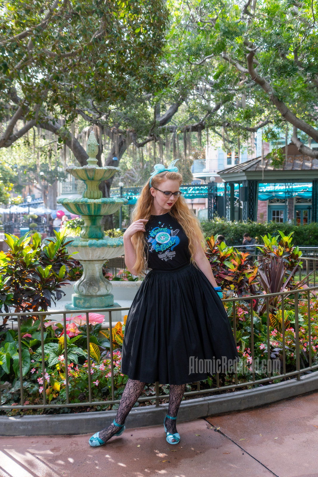 Myself standing in front of a fountain surrounded by trees dripping in Spanish moss, wearing a black t-shirt with Madam Leota (a medium with her head floating in a crystal ball) and music instruments floating about on it, reading "Having a Ball" and a black full skirt.