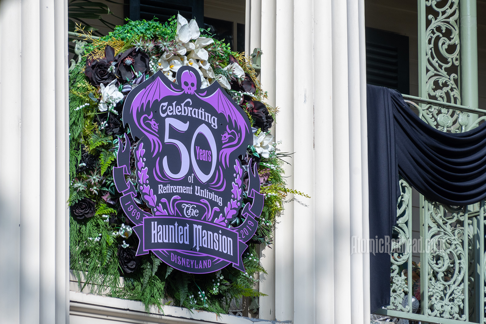 A purple and black sign hangs on the balcony of the Haunted Mansion reading "Celebrating 50 years of Retirement Unliving" The Haunted Mansion 1969 - 2019