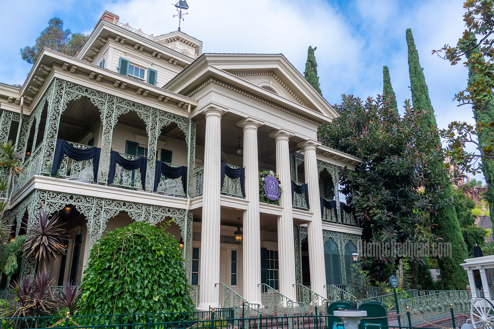 The Haunted Mansion, a stately two story Greek Revival, antebellum mansion with white pillars in center and curling green wrought iron around the side.