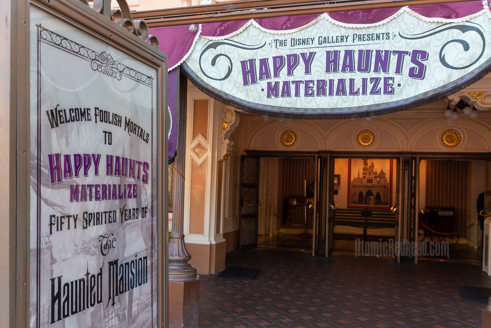 A sign in the left foreground reads "Welcome Foolish Mortals to the Happy Haunts Materialize Fifty Spirited Years of the Haunted Mansion" in the background a banner reads "The Disney Gallery Presents Happy Haunts Materialize"
