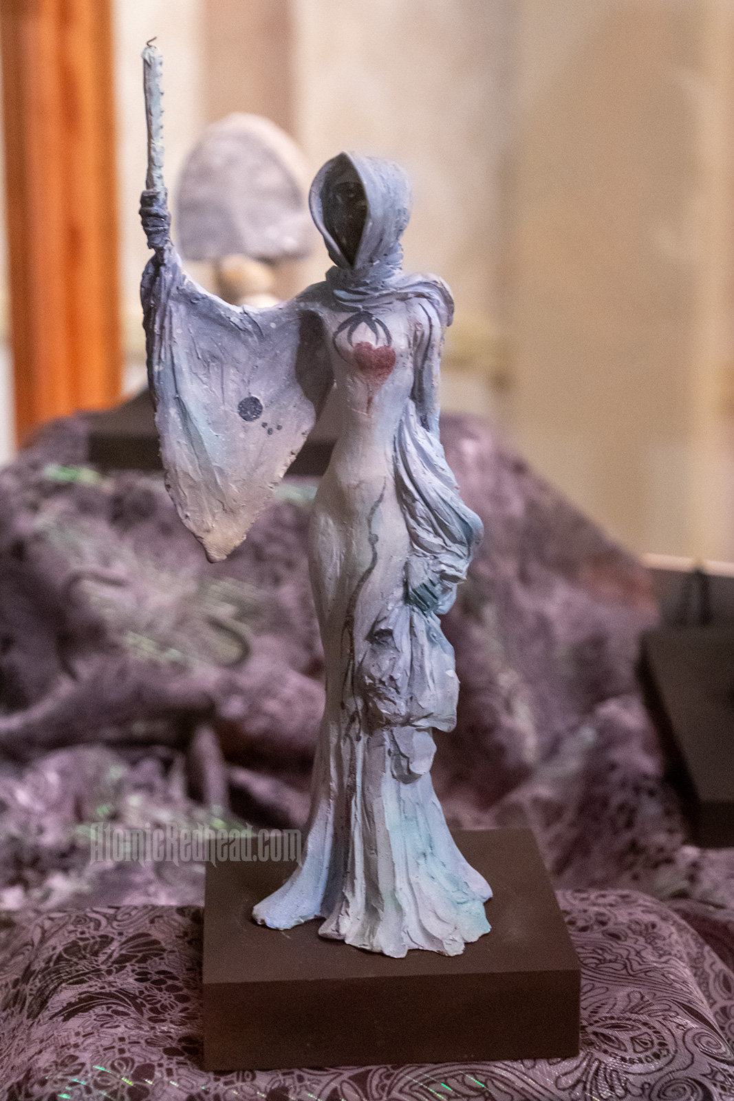 An ghastly sculpture of a woman in a hooded gown with long flowing sleeves. Her face is dark, but her heart is show beating from under her dress, she holds a candle. 