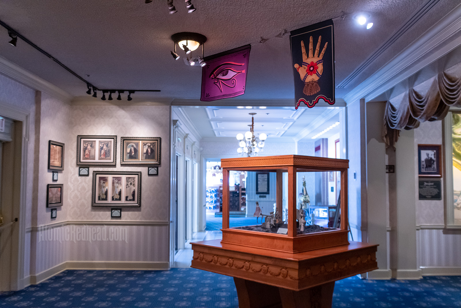Overall image of the gallery, featuring concept art on the wall, a display case with small sculptures within, above hang two banners, one of the Eye of Ra, the other features a hand wrapped in ribbon with a rose in the center.