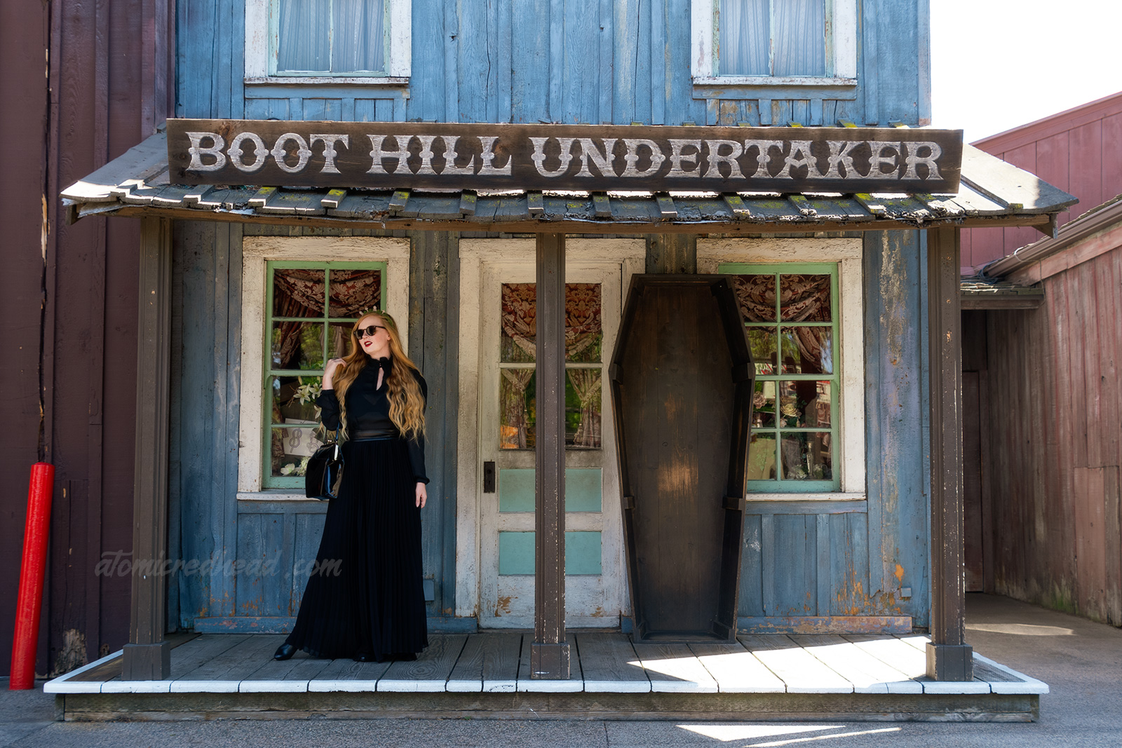 Myself, wearing a black long sleeve high collar blouse, and a long black skirt, holding a black coffin shaped purse, and a tiara made of small tombstones, standing in front the the Boot Hill Undertaker, a western style building with a coffin standing out front.