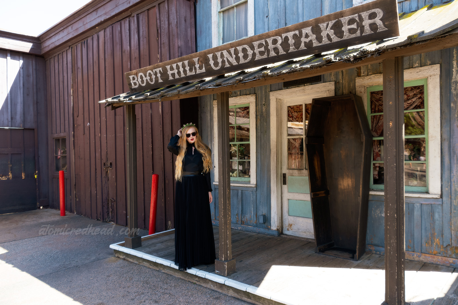 Myself, wearing a black long sleeve high collar blouse, and a long black skirt, and a tiara made of small tombstones, standing in front the the Boot Hill Undertaker, a western style building with a coffin standing out front.