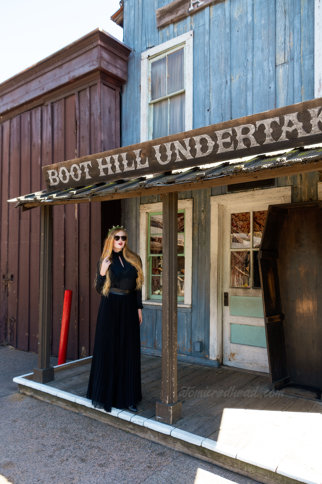 Myself, wearing a black long sleeve high collar blouse, and a long black skirt, and a tiara made of small tombstones, standing in front the the Boot Hill Undertaker, a western style building with a coffin standing out front.