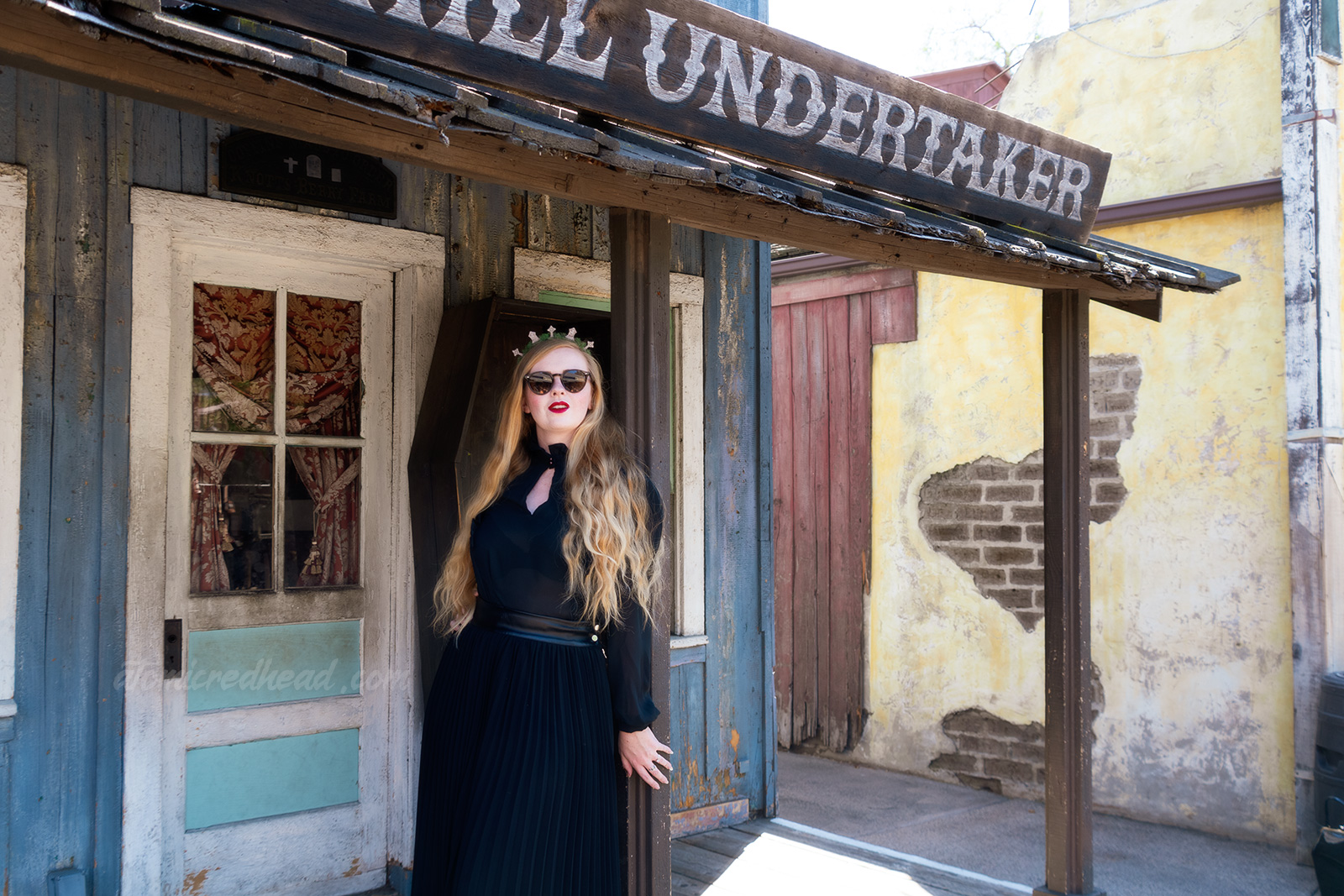 Myself, wearing a black long sleeve high collar blouse, and a long black skirt, and a tiara made of small tombstones, standing in front the the Boot Hill Undertaker, a western style building with a coffin standing out front.