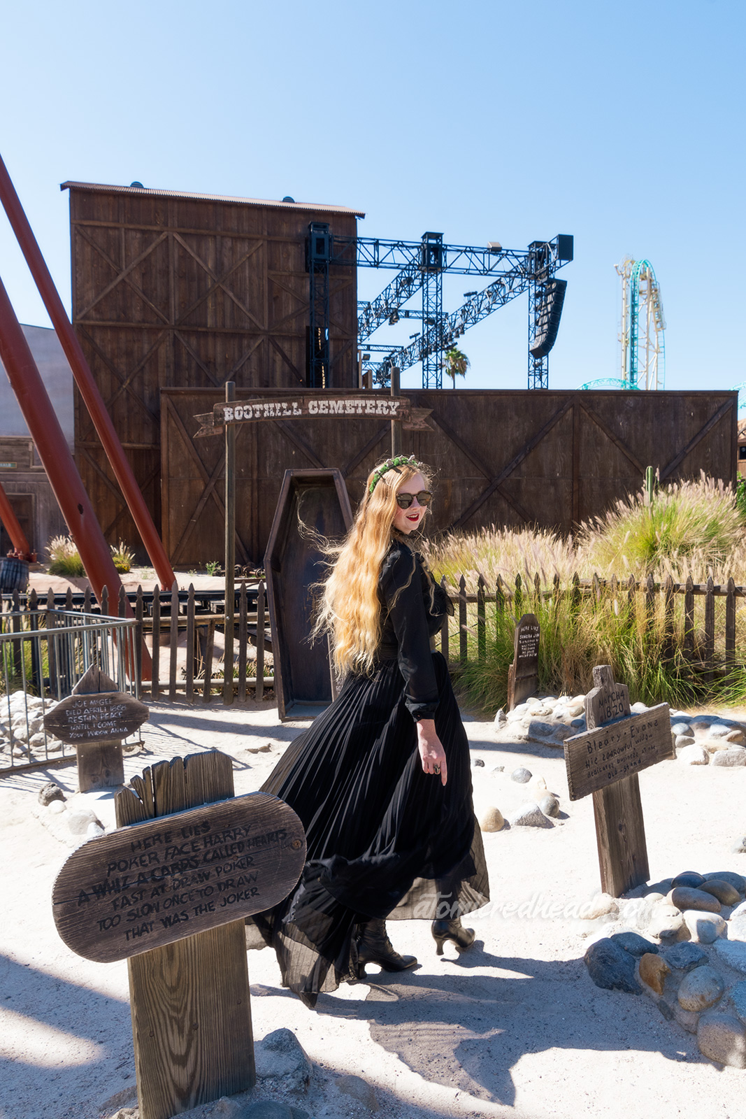 Myself, wearing a black long sleeve high collar blouse, and a long black skirt, holding a black coffin shaped purse, and a tiara made of small tombstones, walking through the Boot Hill cemetery, which features wooden tombstones.