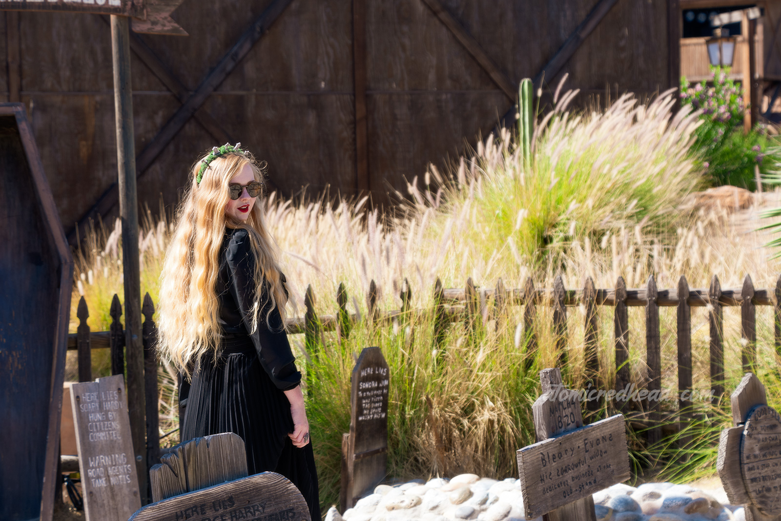 Myself, wearing a black long sleeve high collar blouse, and a long black skirt, holding a black coffin shaped purse, and a tiara made of small tombstones, walking through the Boot Hill cemetery, which features wooden tombstones.