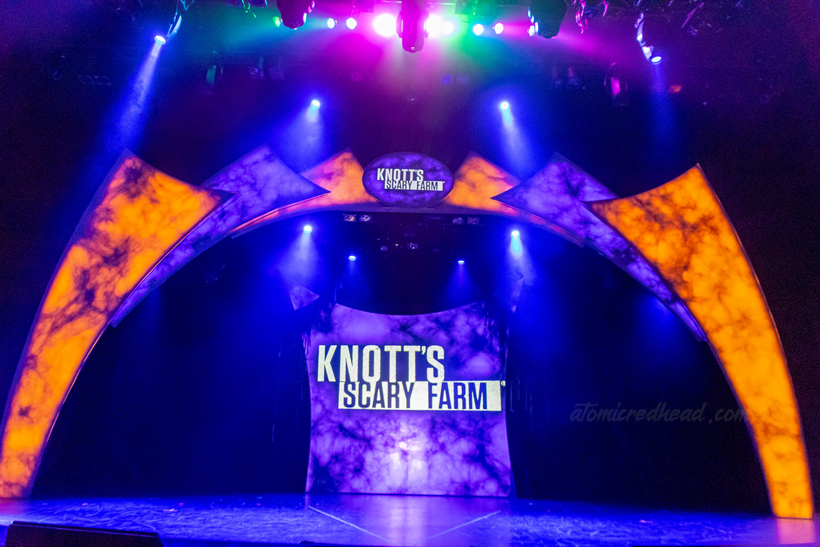The stage for the announcement for Knott's Scary Farm, lit in eerie orange and purple lights. Text reading "Knott's Scary Farm" in white letters is on a screen at the back of the stage and a small sig at the top of the stage.