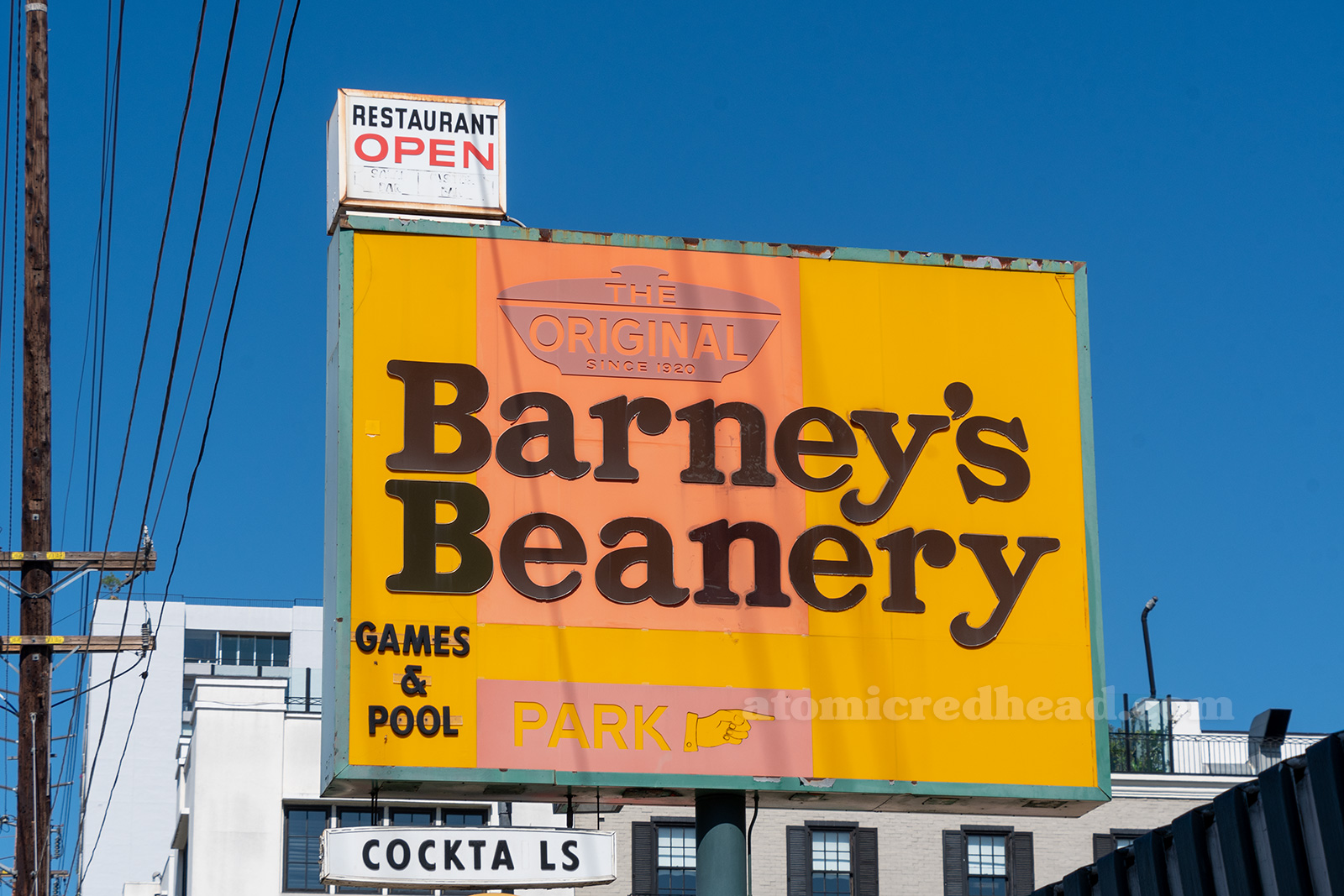 Close-up of the sign, a small white sign sits on top of a larger sign. The small sign reads "Restaurant Open" the larger sign reads "The Original Barney's Beanery Games & Pool" another smaller white sign reads "Cocktails"