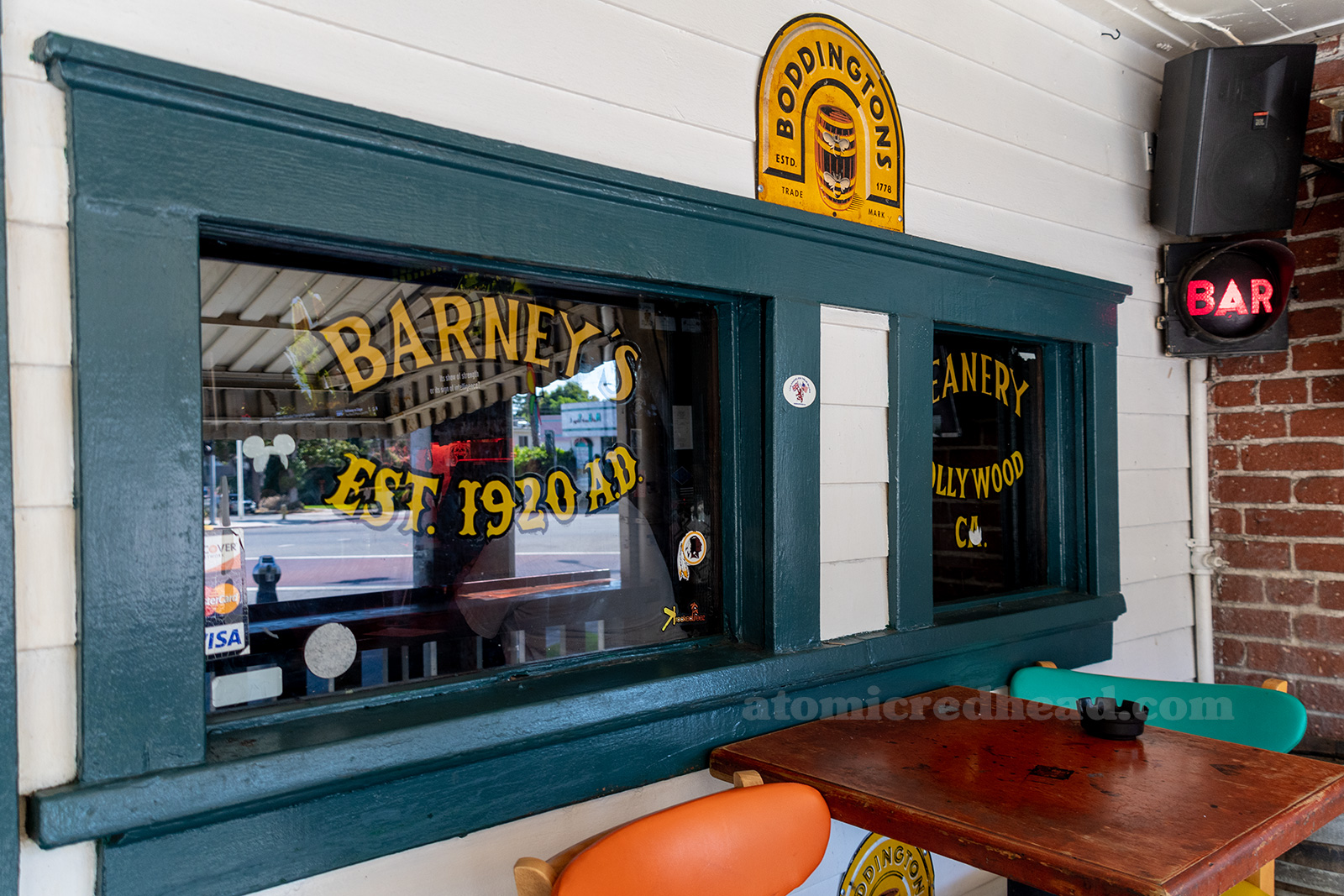The frame of a window is painted green, the rest of the building white, the text on the window reads "Barney's Beanery Est. 1920 AD Hollywood California"