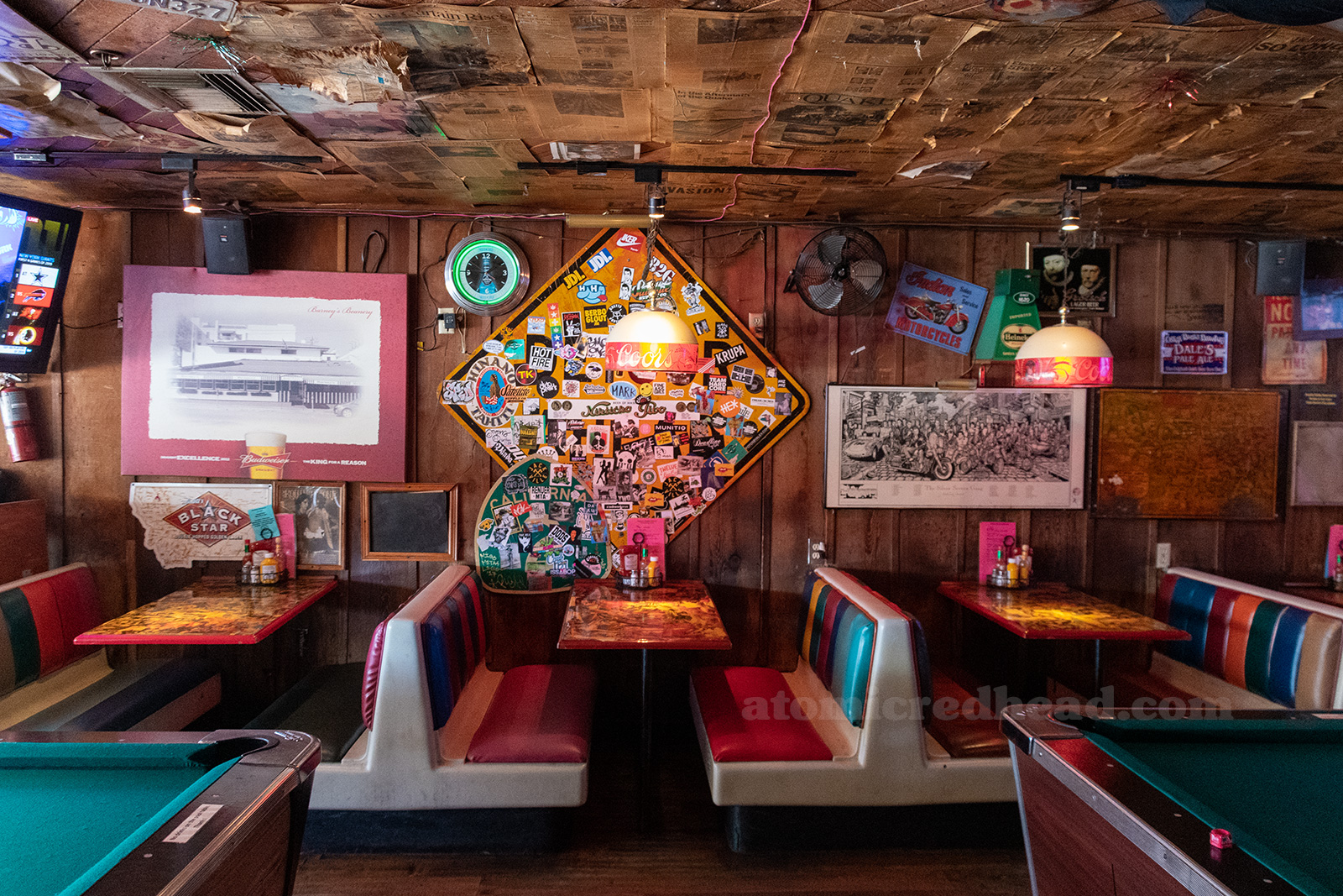 Interior of Barney's, newspapers cover the ceiling, artwork, signs, and other odds and ends hang from wood paneled walls. Multi color booths sit against the wall, pool tables are just off frame.