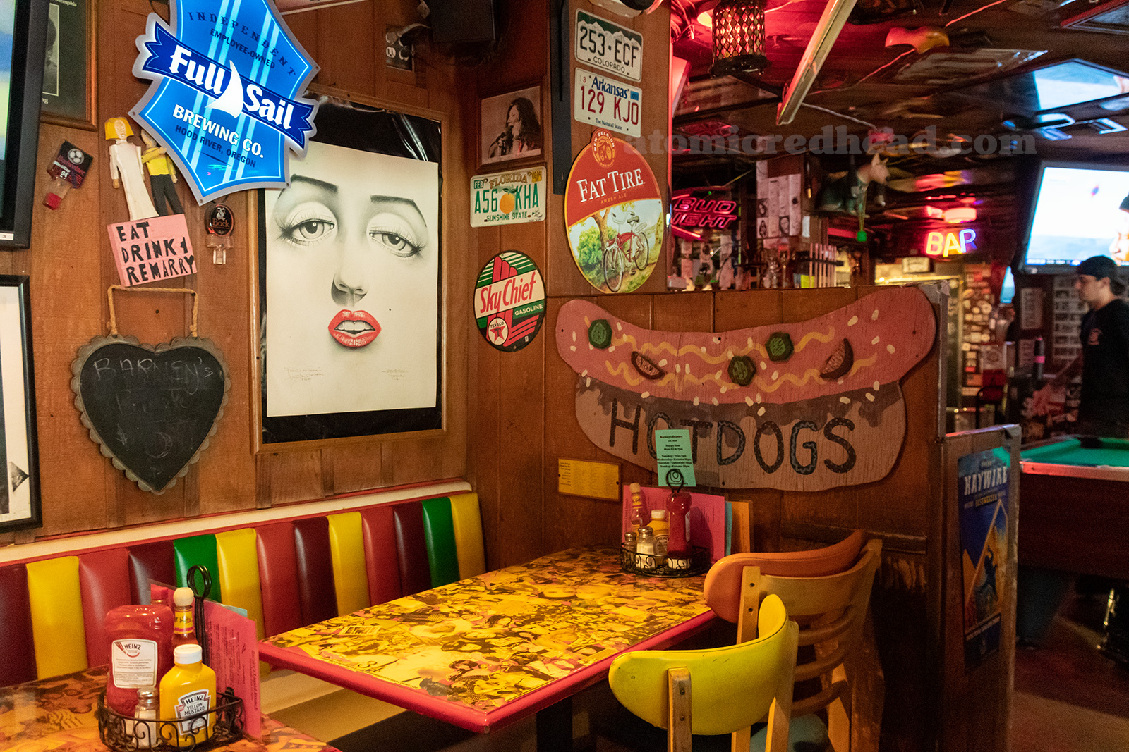 Interior of Barney's, newspapers cover the ceiling, artwork, signs, and other odds and ends hang from wood paneled walls. Multi color booths sit against the wall, pool tables are just off frame.
