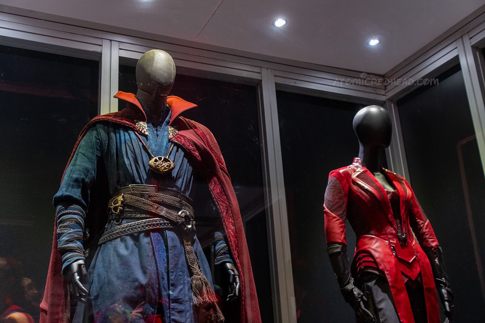 Dr. Strange and Scarlet Witch's costumes from Avengers. Dr. Strange wears a navy ensemble with multiple brown leather belts, and a red cape, his iconic eye shaped necklace hangs. Scarlet Witch wears a tight fitting red ensemble.