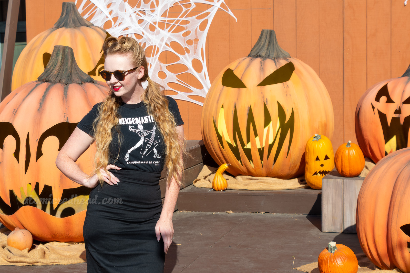 Myself, wearing a black t-shirt with a zombie playing an upright bass shaped like a coffin, and text reading "Nekromatix Coffinbass is Cool" and a black pencil skirt, standing in front of a stage with massive jack o'lanterns.