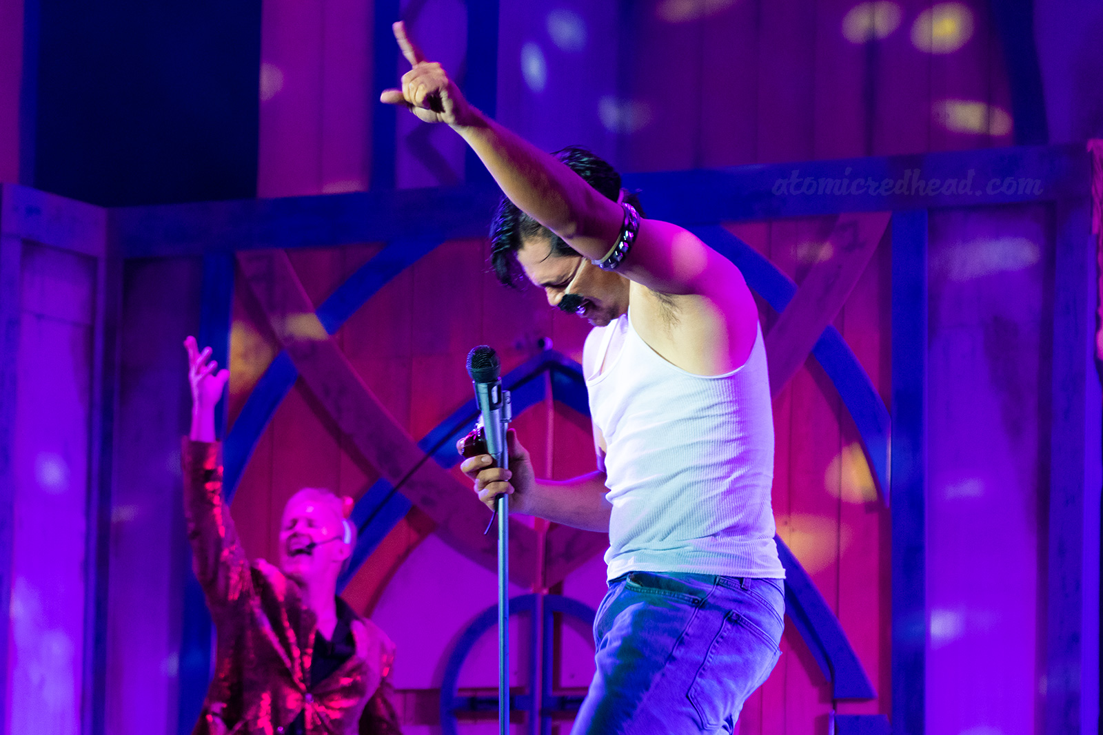 A man dressed as Freddie Mercury sings into a microphone. Behind him a man dressed as the devil mimics him.