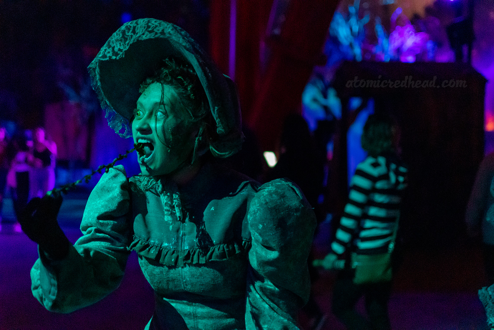 A woman in grey Victorian dress pulls a string of leaves from her mouth, as the victims of the Lake wonder through the Scare Zone of Forsaken Lake.