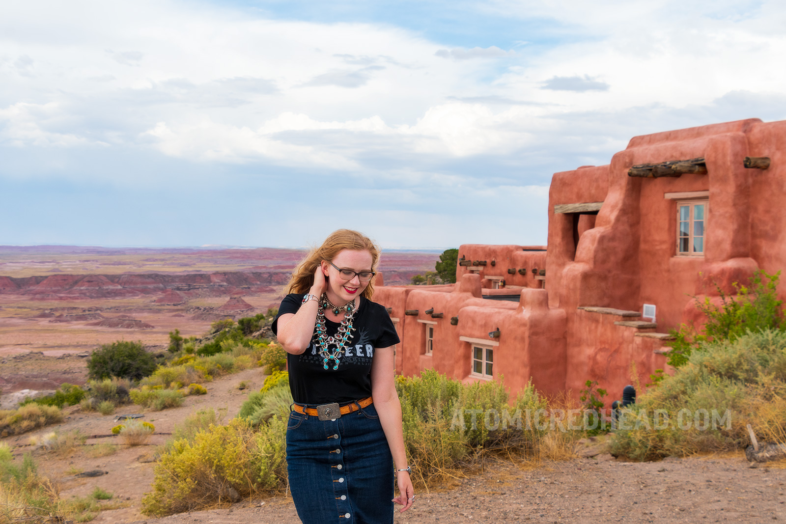 Myself, wearing several turquoise and silver necklaces, a black t-shirt reading "Pioneer" in white letters, and a jean skirt, standing in front of the Painted Desert Inn.