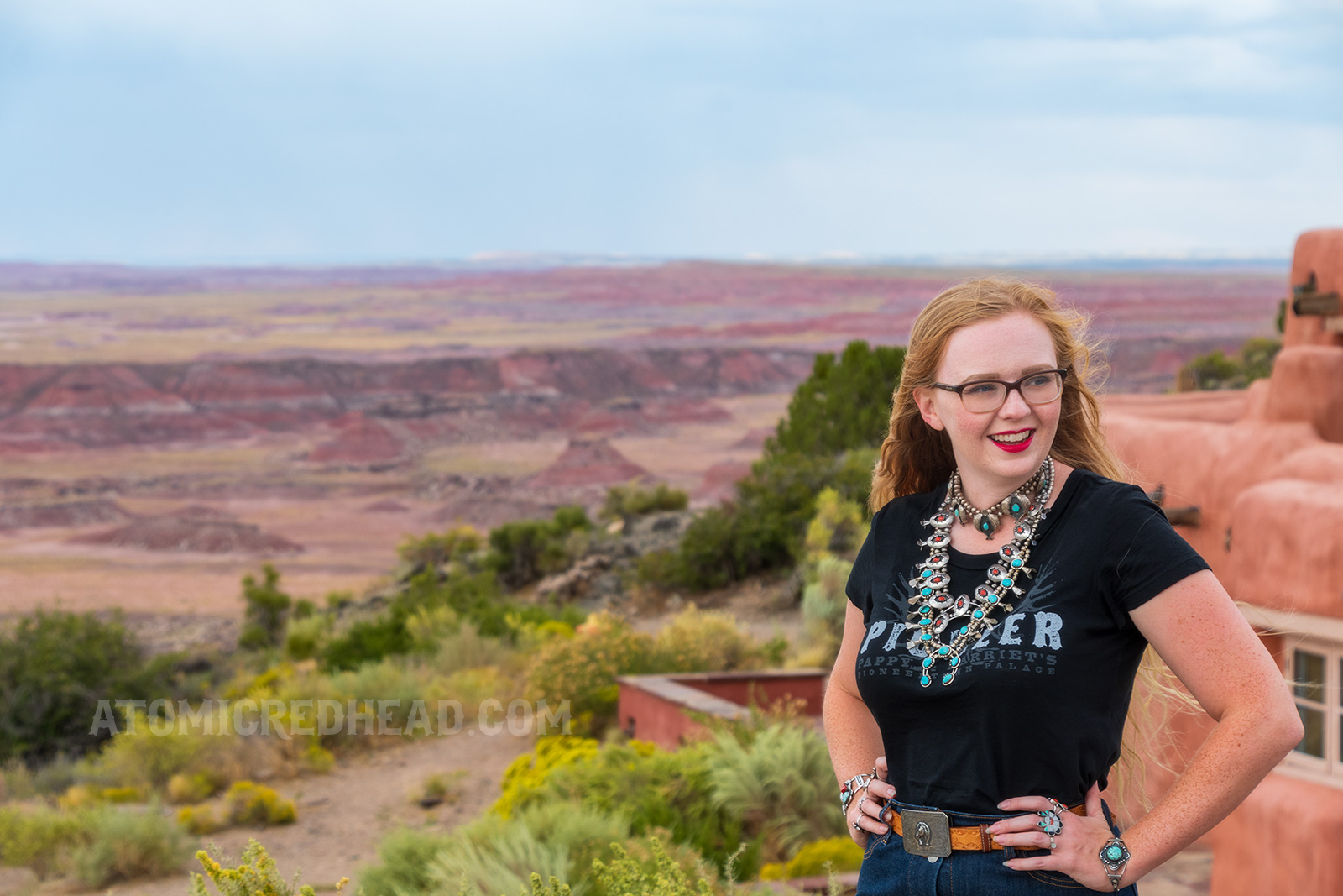Myself, wearing several turquoise and silver necklaces, a black t-shirt reading "Pioneer" in white letters, and a jean skirt, standing in front of the Painted Desert Inn.