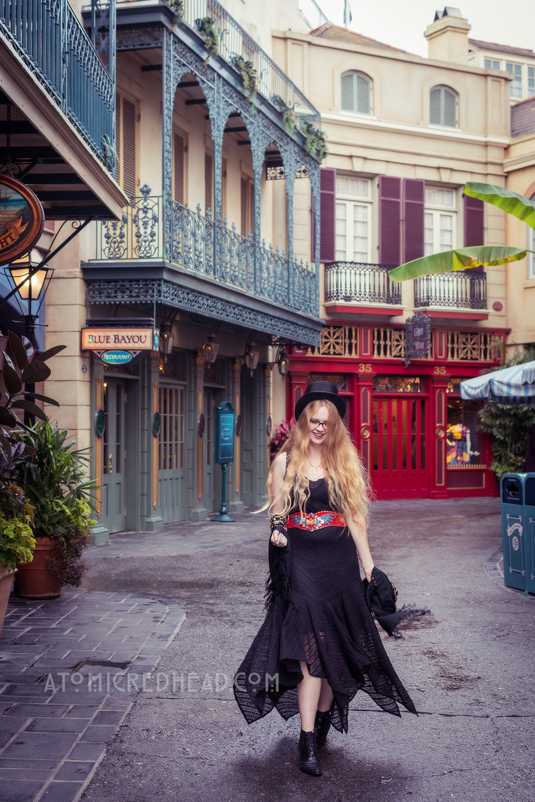 Myself below the green curling wrought iron balconies of New Orleans Square. Wearing a black top hat, black lace dress, with red, blue, and gold belt featuring an eagle, and short black boots.