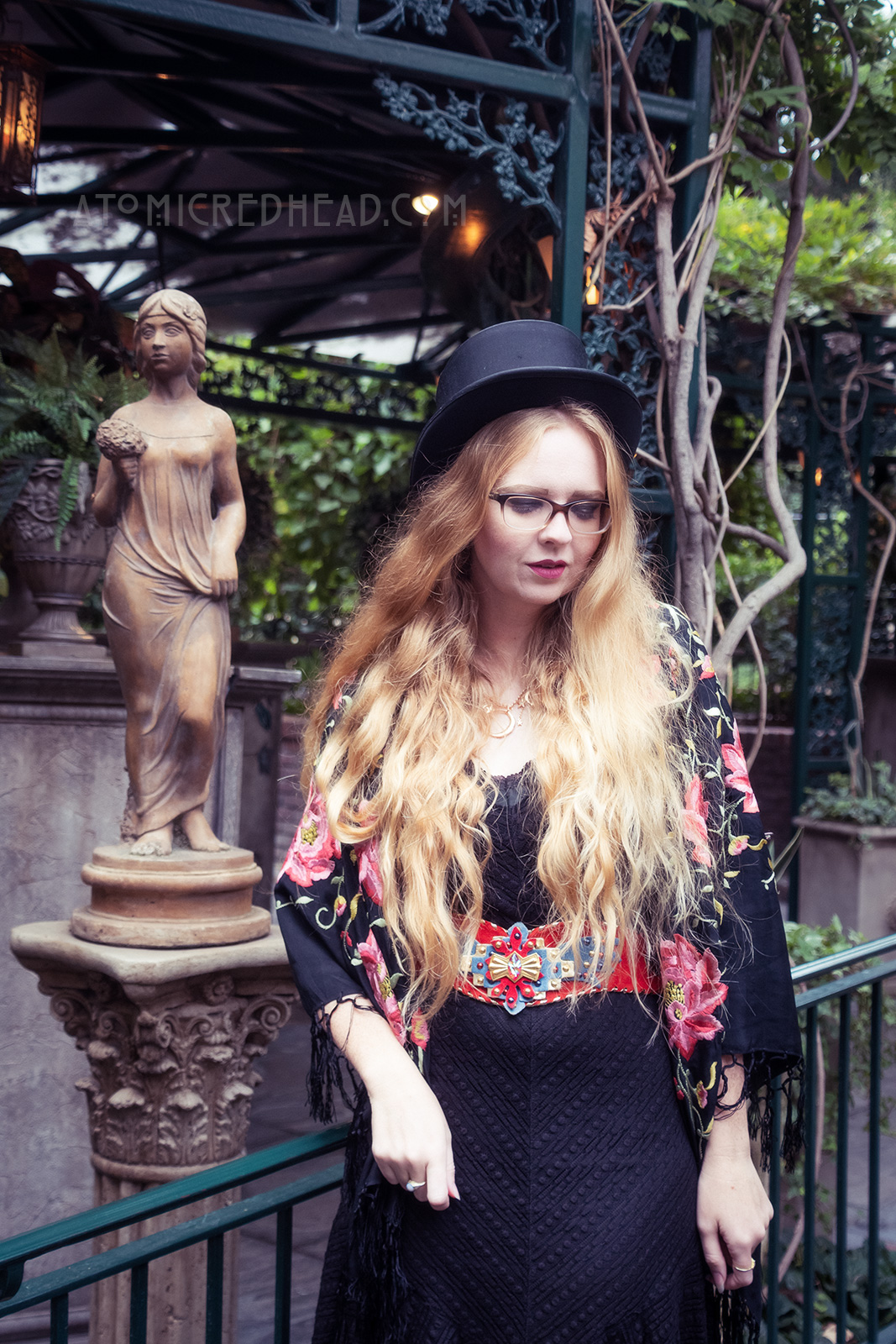 Myself standing near a statue of a woman, surrounded by leafy trees, wearing a black top hat, black lace dress, with red, blue, and gold belt featuring an eagle, a black shawl with pink flowers embroidered on it around my shoulders, and short black boots.