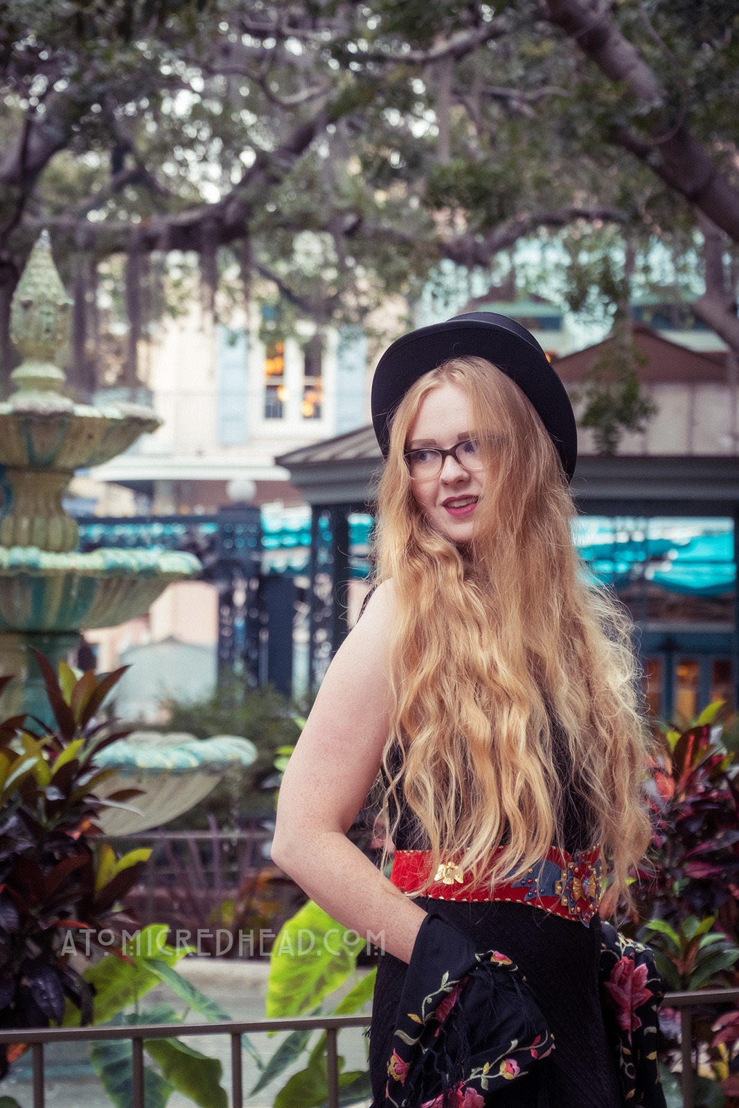 Myself standing near a fountain under trees of Spanish moss, wearing a black top hat, black lace dress, with red, blue, and gold belt featuring an eagle, a black shawl with pink flowers embroidered on it around my shoulders, and short black boots.