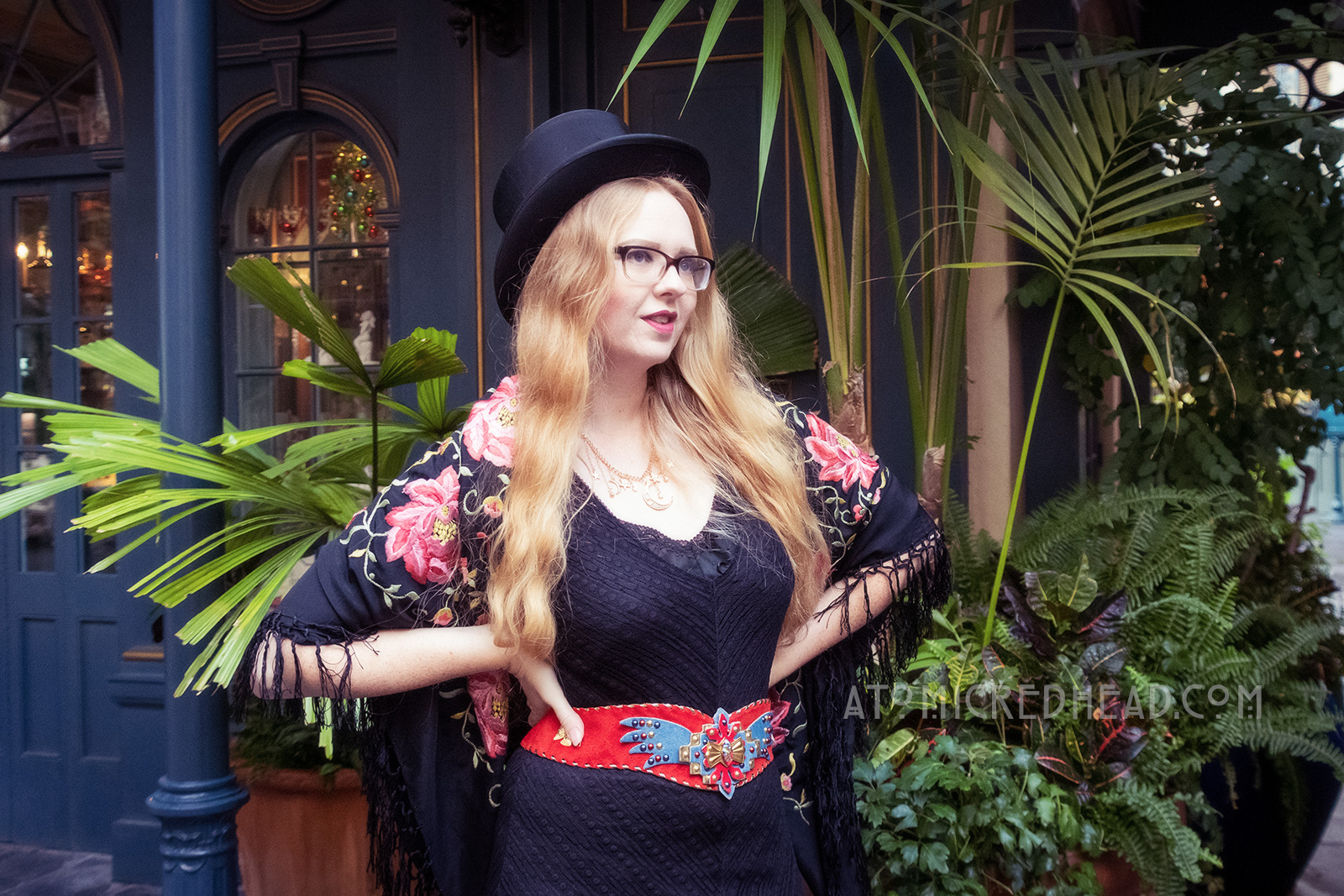 Myself standing in front of one of New Orleans Square's shops, wearing a black top hat, black lace dress, with red, blue, and gold belt featuring an eagle, a black shawl with pink flowers embroidered on it around my shoulders, and short black boots.