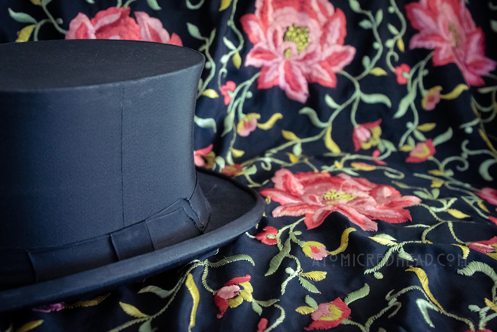 Close-up, a black top hat sits on a black shawl with pink flowers embroidered on it.