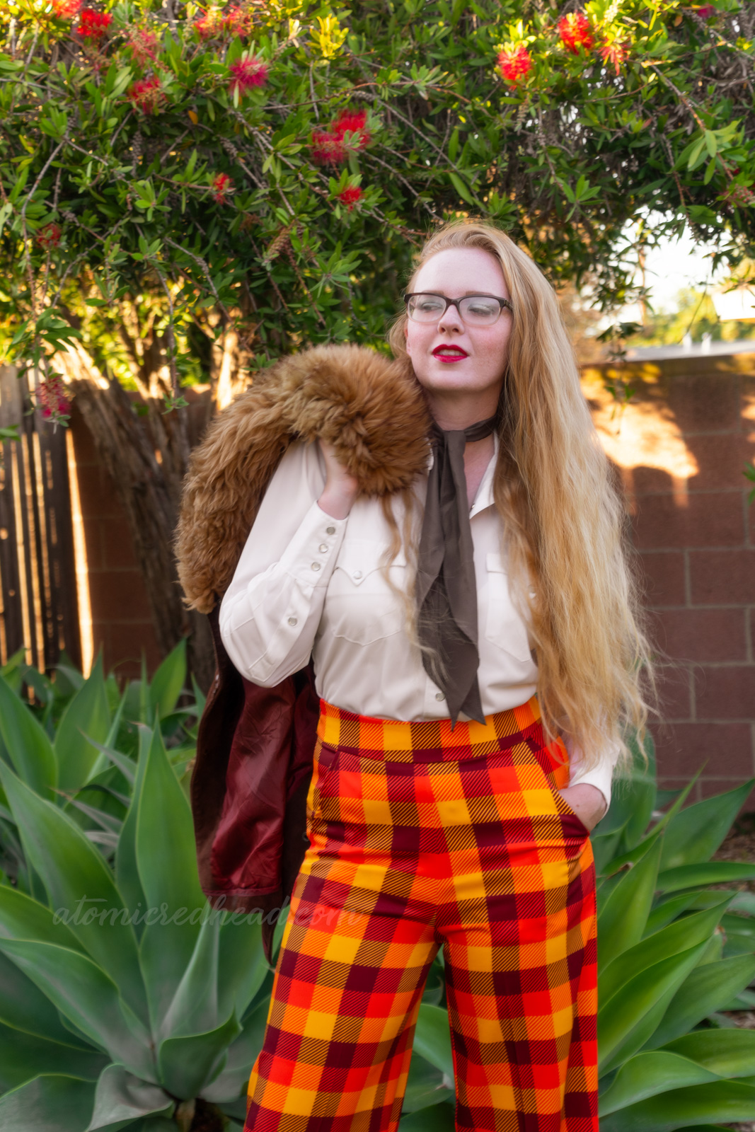 Myself, wearing a white western wear top, brown scarf, and yellow, orange, and brown plaid pants, a brown jacket is thrown over my shoulder.