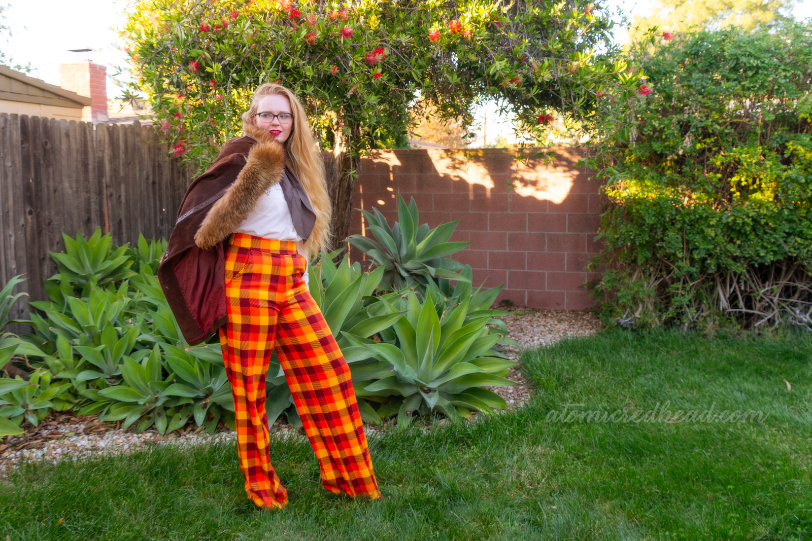 Myself, wearing a white western wear top, brown scarf, and yellow, orange, and brown plaid pants, a brown jacket is thrown over my shoulder.