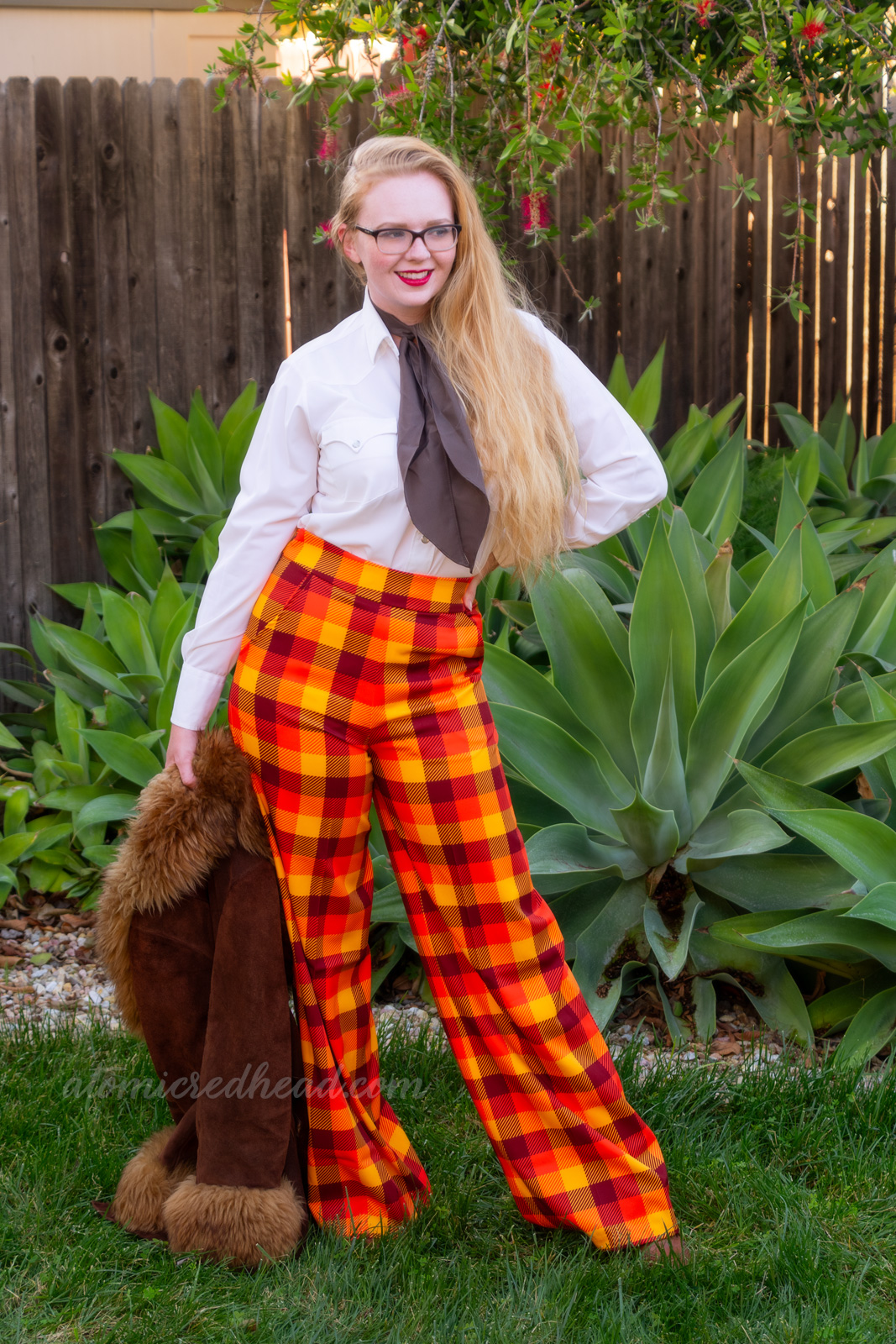Myself, wearing a white western wear top, brown scarf, and yellow, orange, and brown plaid pants, a brown jacket is held in my right hand.