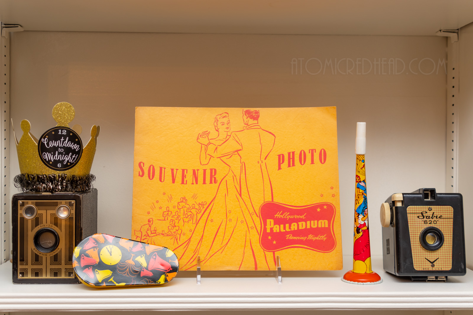 A souvenir photo folder from the Hollywood Palladium features a dancing couple, it is flanked by two black and gold cameras, vintage noise makers, and a gold crown reading "Countdown to Midnight"