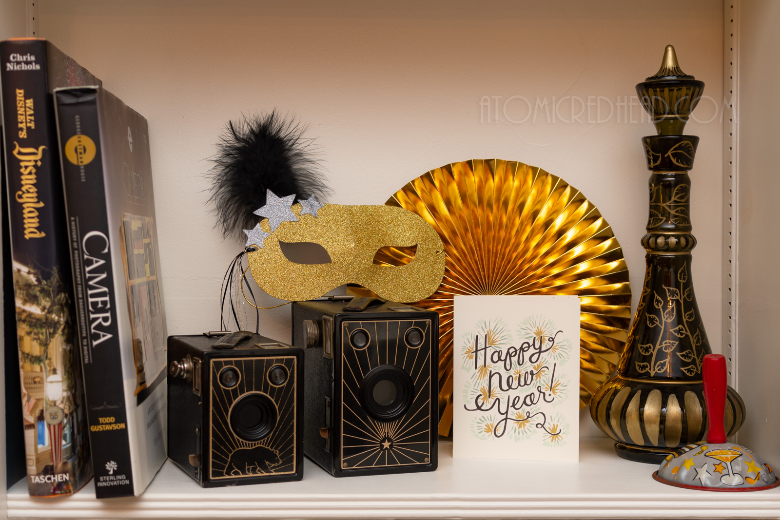 A gold glitter mask sits on top of two black and gold cameras, a card reading "Happy New Year" sits next to the cameras.