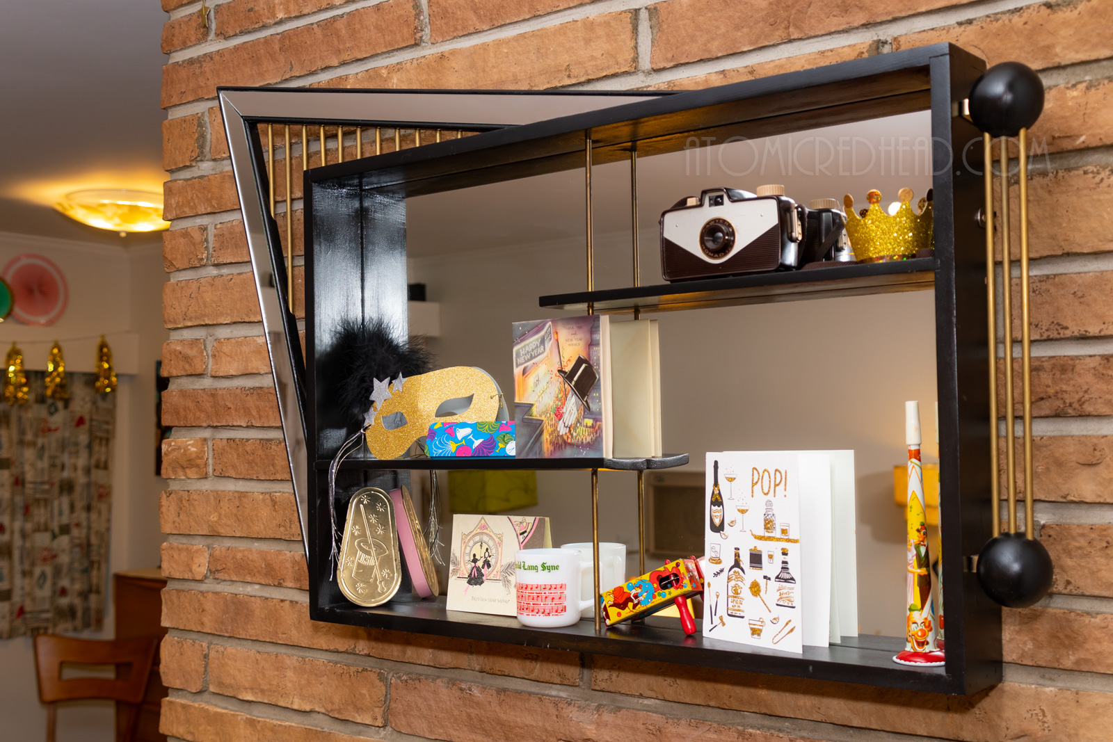 A black and gold mirrored shadowbox hangs on our brink fireplace. Inside sit vintage noise makers, a gold glitter mask, and vintage New Years cards.