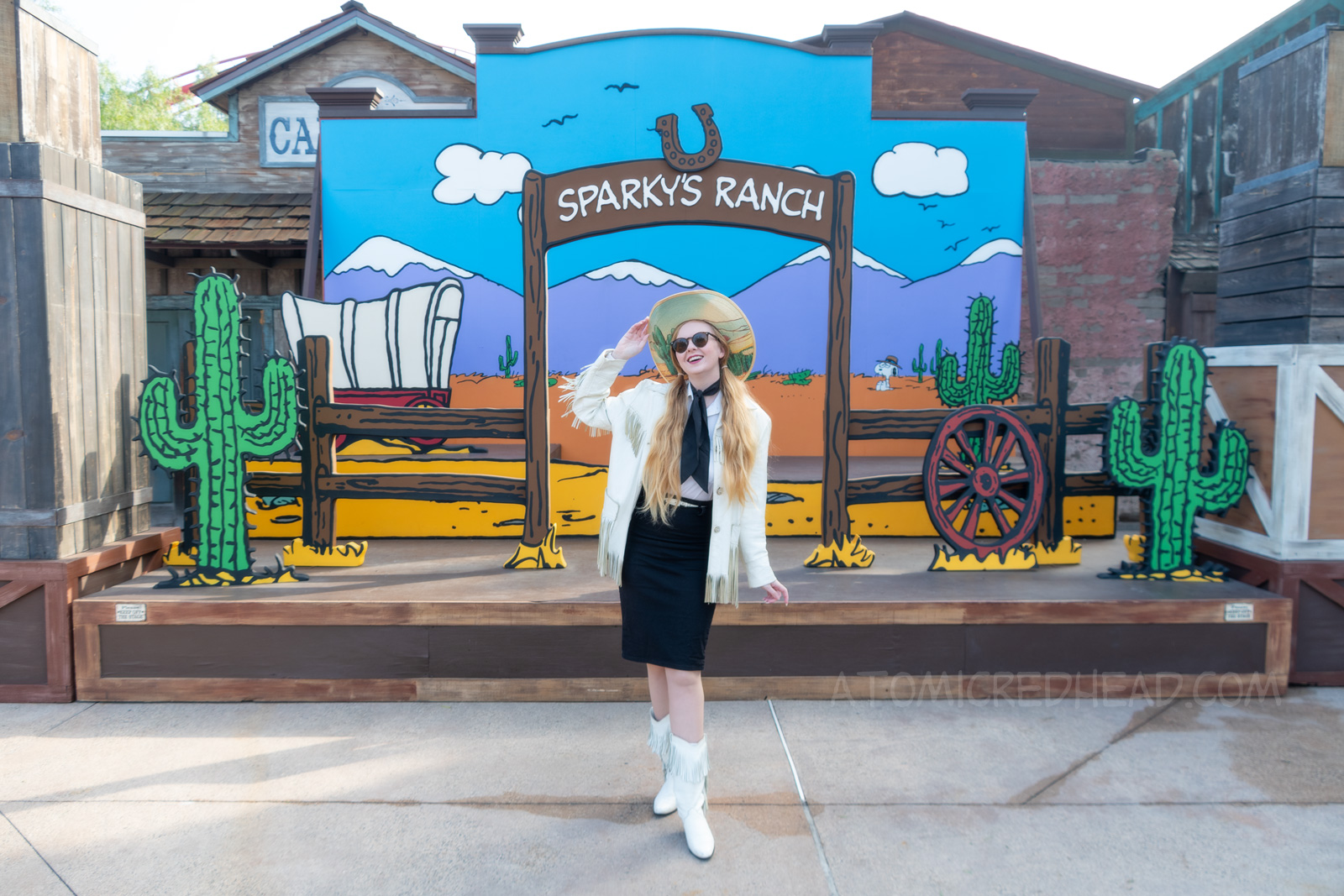 Myself standing in front of a western backdrop done in the style of Peanuts creator Charles M. Schulz. I'm wearing a cowboy hat with a cactus print on the brim, a white fringe leather jacket, a white western wear shirt, a black scarf, black skirt, and white fringe leather boots.
