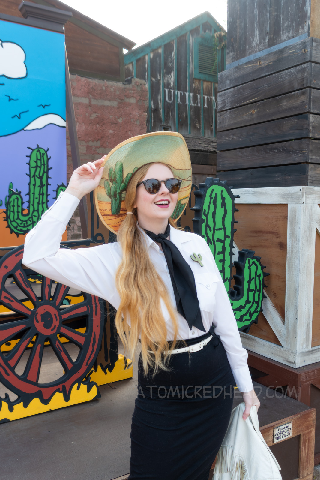 Myself standing in front of a western backdrop done in the style of Peanuts creator Charles M. Schulz. I'm wearing a cowboy hat with a cactus print on the brim, a white fringe leather jacket, a white western wear shirt, a black scarf, black skirt, and white fringe leather boots.