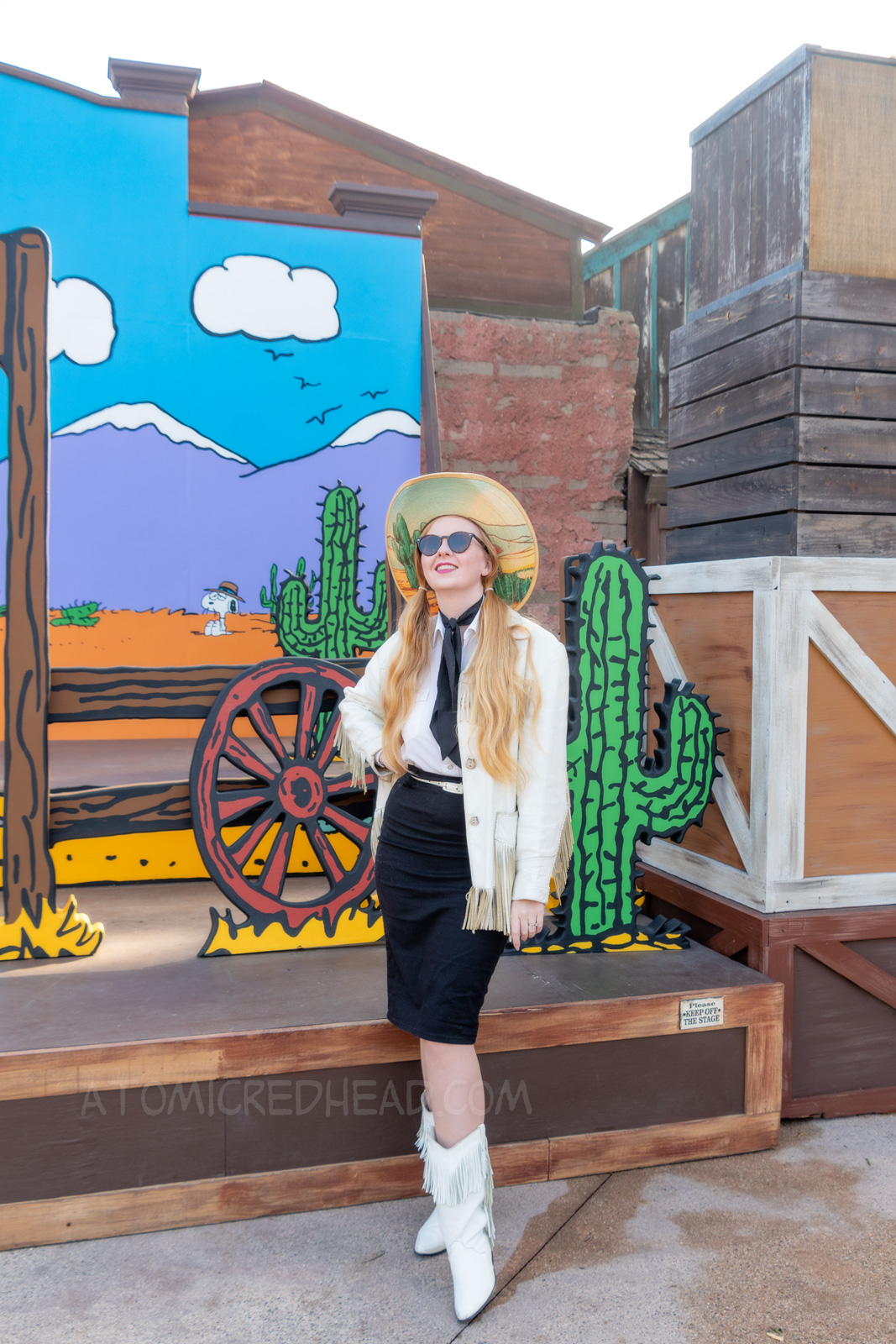 Myself standing in front of a western backdrop done in the style of Peanuts creator Charles M. Schulz. I'm wearing a cowboy hat with a cactus print on the brim, a white fringe leather jacket, a white western wear shirt, a black scarf, black skirt, and white fringe leather boots.