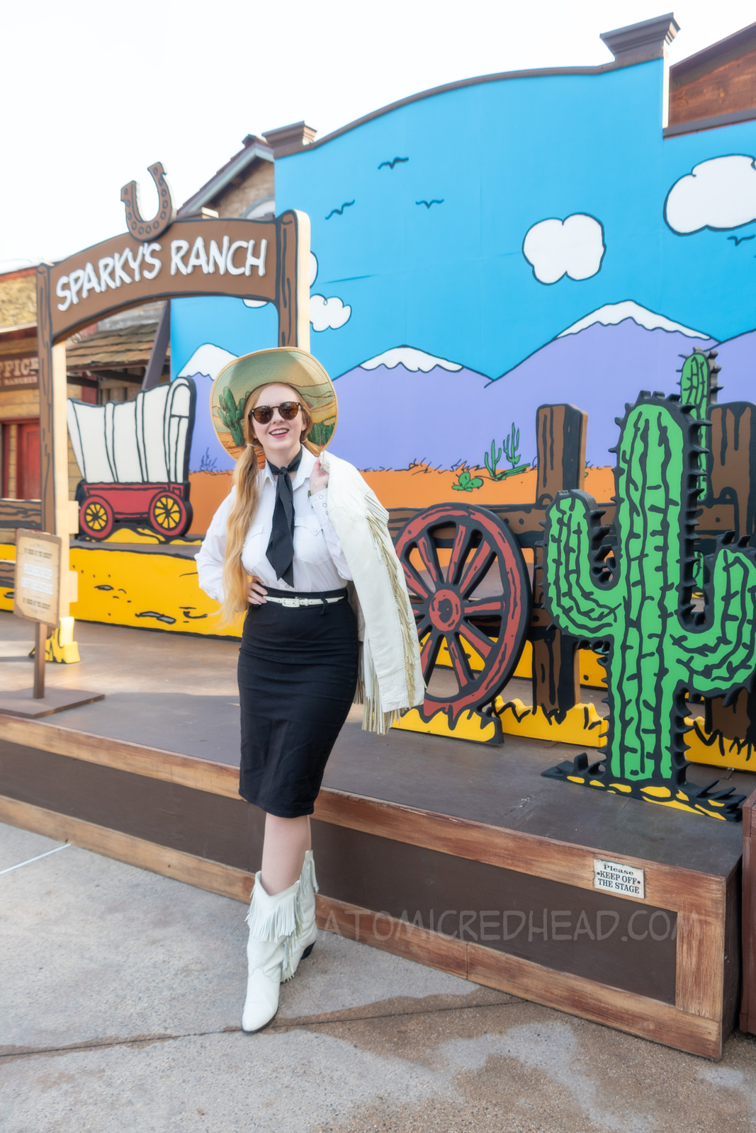 Myself standing in front of a western backdrop done in the style of Peanuts creator Charles M. Schulz. I'm wearing a cowboy hat with a cactus print on the brim, a white fringe leather jacket, a white western wear shirt, a black scarf, black skirt, and white fringe leather boots.