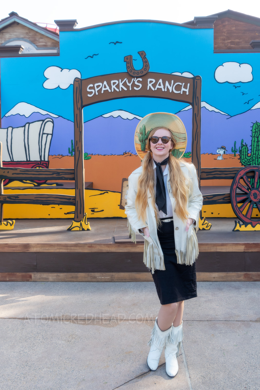 Myself standing in front of a western backdrop done in the style of Peanuts creator Charles M. Schulz. I'm wearing a cowboy hat with a cactus print on the brim, a white fringe leather jacket, a white western wear shirt, a black scarf, black skirt, and white fringe leather boots.