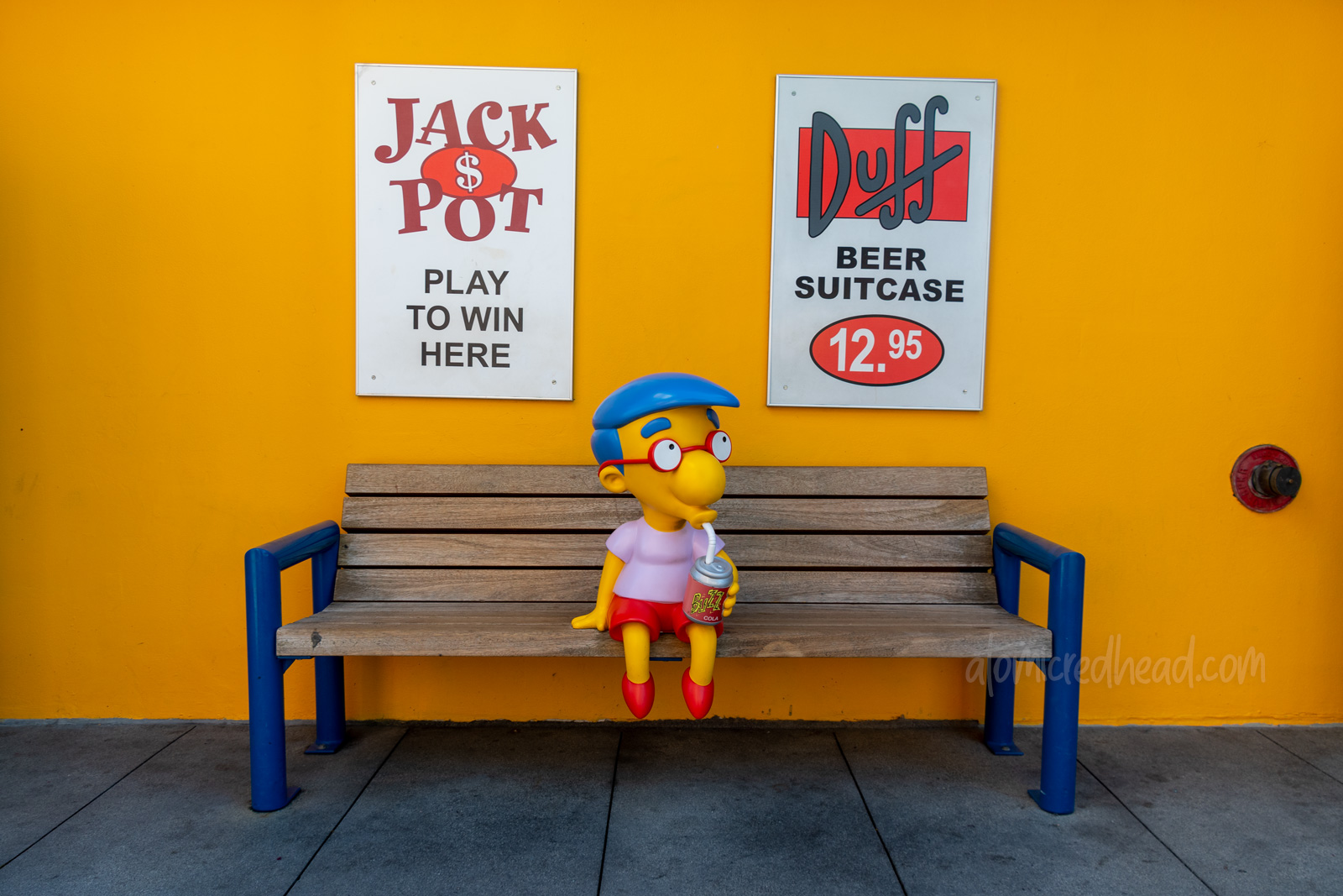 Statue of Milhouse sits on a bench drinking a Buzz Cola.