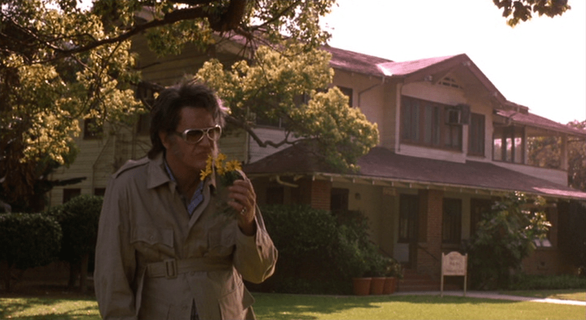 Screencap from Bubba Ho-Tep: An older Elvis stands in front of a two story craftsman house, holding a flower.
