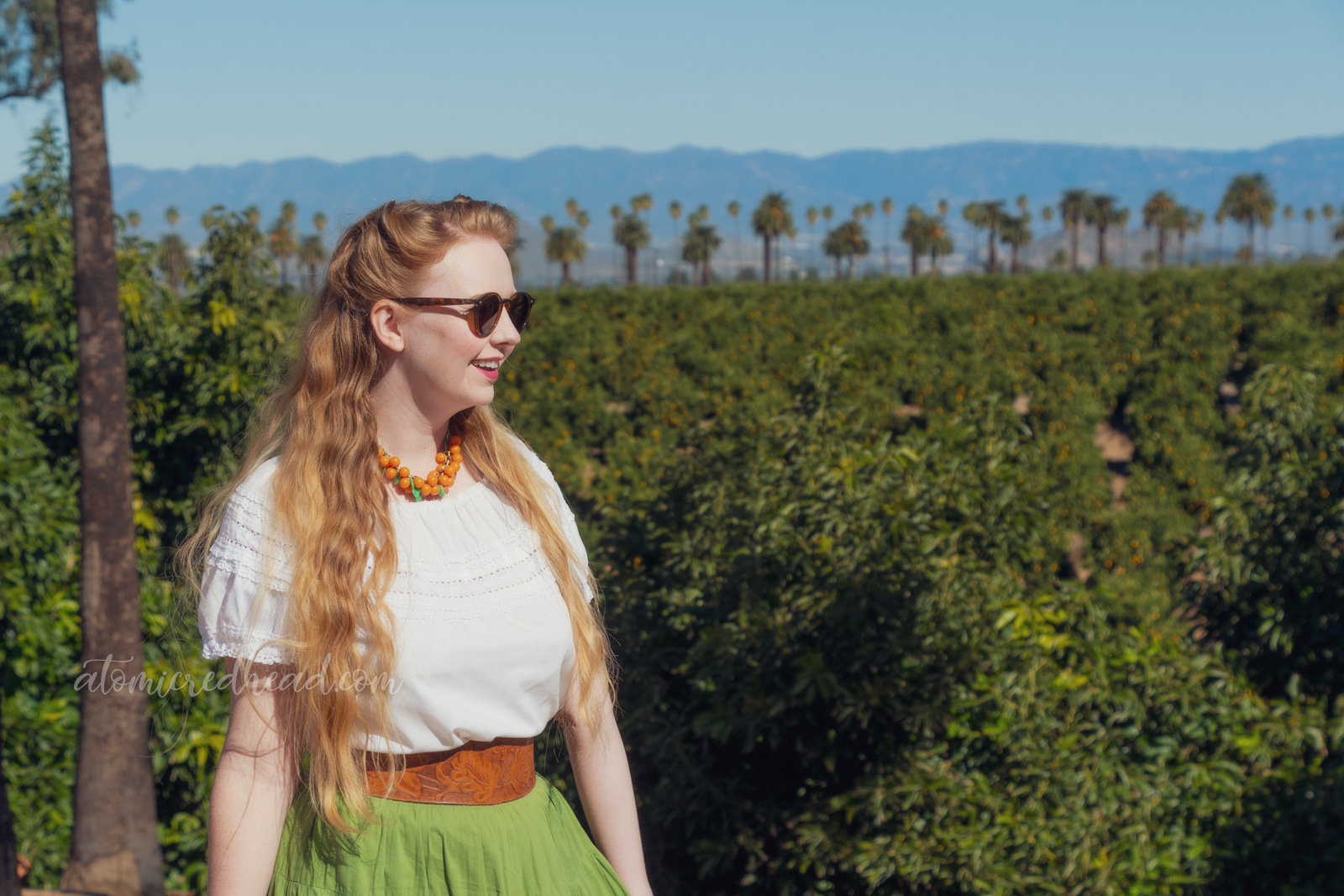 Myself, wearing a necklace of Bakelite oranges and leaves, a white peasant top, and a green skirt with orange, gold, and dark green rick-rack, standing among orange trees.