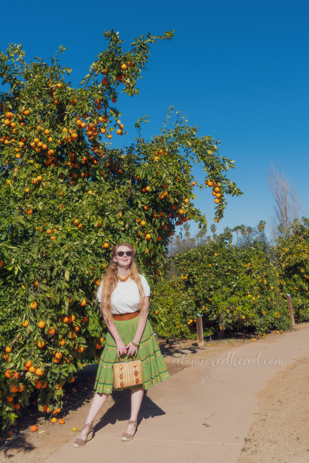Myself, wearing a necklace of Bakelite oranges and leaves, a white peasant top, and a green skirt with orange, gold, and dark green rick-rack, standing among orange trees.